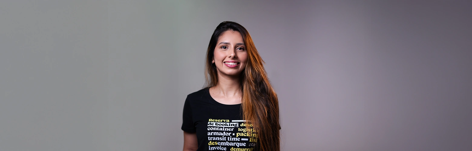 Uma mulher de cabelos castanhos longos está em pé em frente a um fundo cinza liso, sorrindo e vestindo uma camiseta preta com texto em amarelo e branco com palavras relacionadas a transporte, logística e agenciamento de cargas.